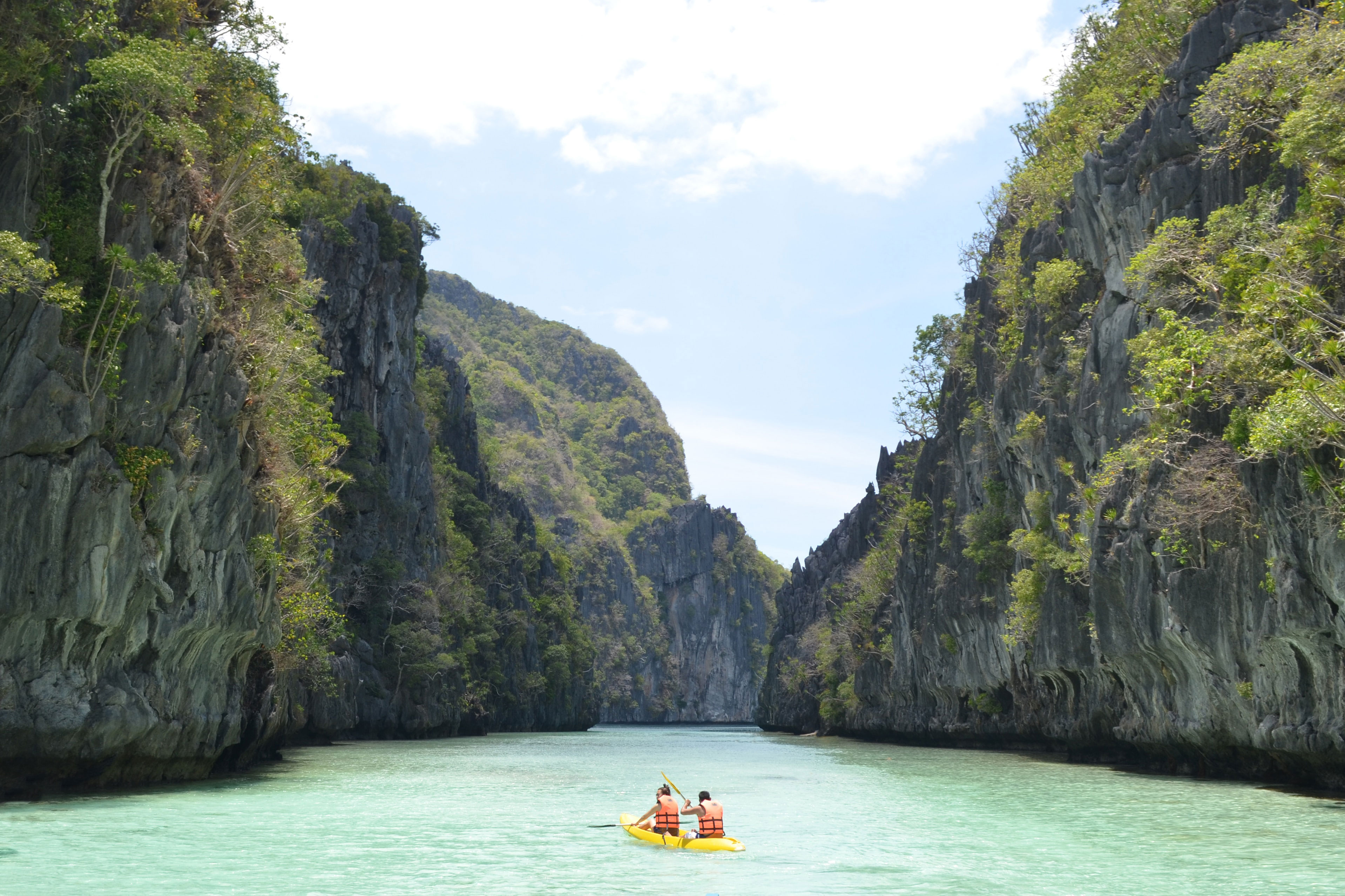 Kayak with two people in it on really clear blue water, surrounded by high cliffs with green vegetation in the Philippines
